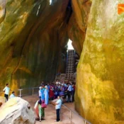 Tourists gather between towering yellowish rock formations at Edakkal Cave in Wayanad, where a staircase leads up in the background.