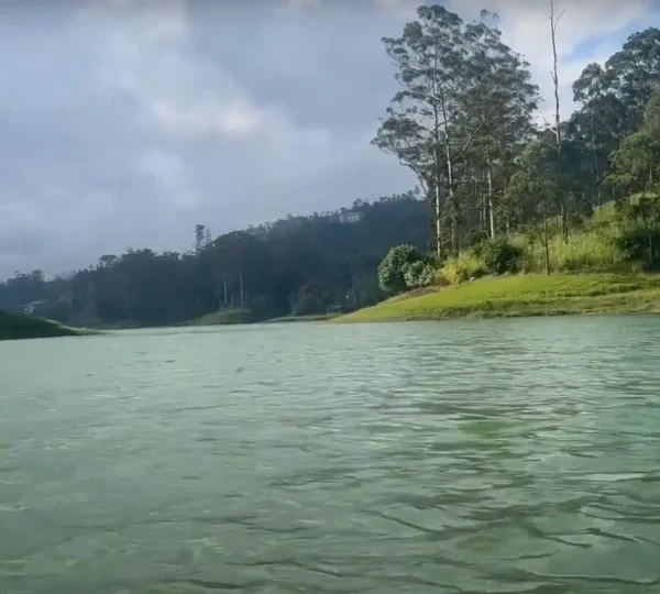 Kundala Lake lies nestled among lush green hills and tall trees under a cloudy sky. Its serene waters mirror the surrounding greenery, crafting a tranquil and picturesque landscape.