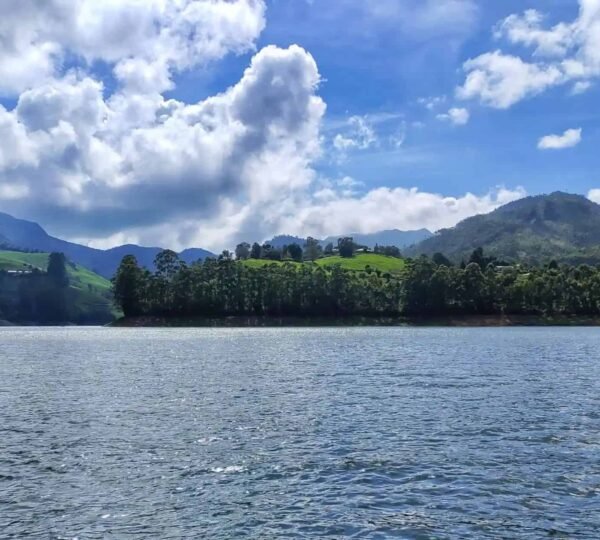 A serene landscape featuring a large lake with calm water in the foreground. Behind the lake, lush green mountains rise under a vibrant blue sky adorned with scattered white clouds.