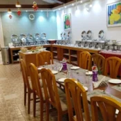 A buffet restaurant among the many renowned restaurants in Munnar, featuring wooden chairs, set dining tables, and a variety of covered serving dishes lined up along the wall under bright lighting.