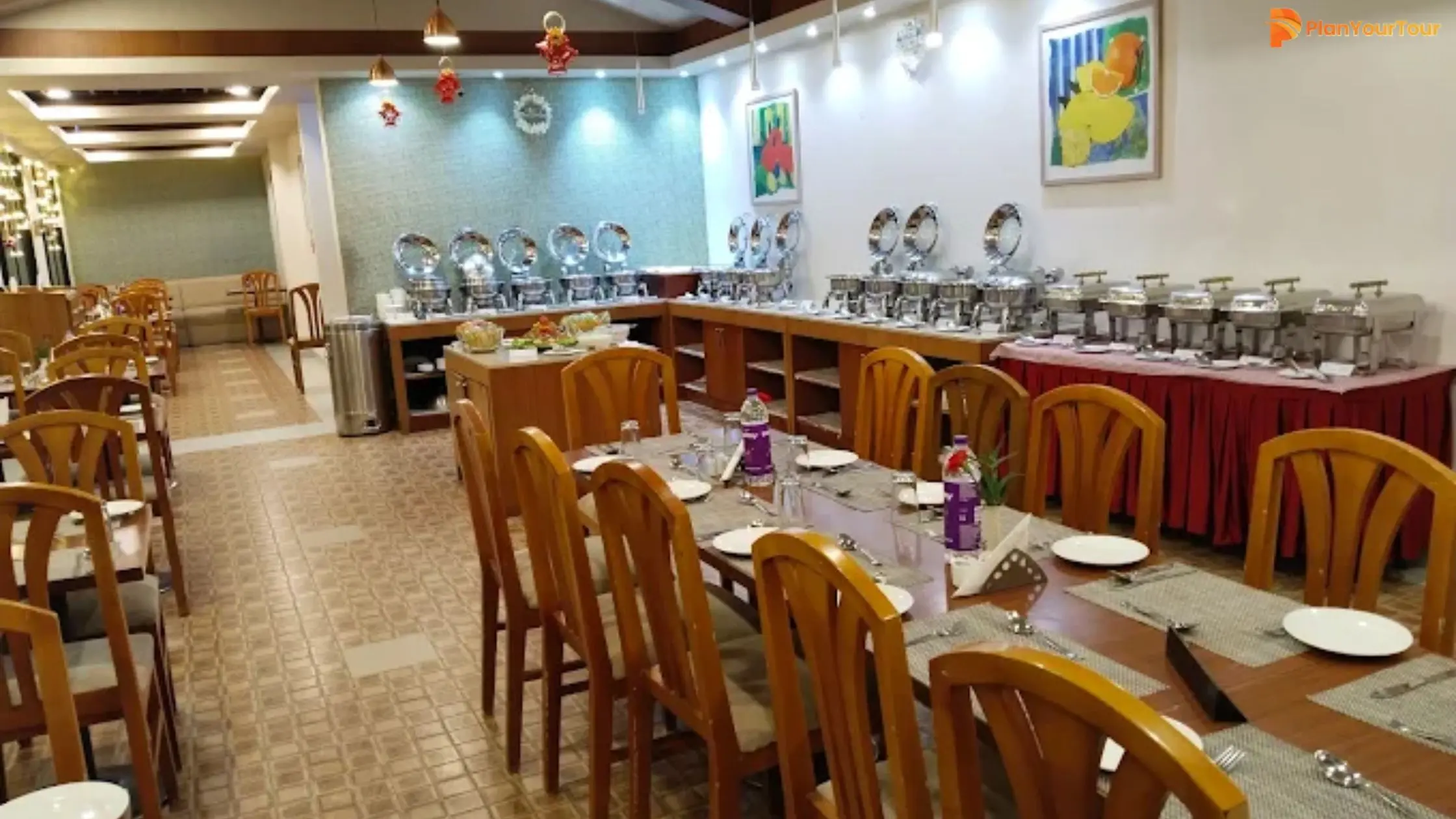A buffet restaurant among the many renowned restaurants in Munnar, featuring wooden chairs, set dining tables, and a variety of covered serving dishes lined up along the wall under bright lighting.